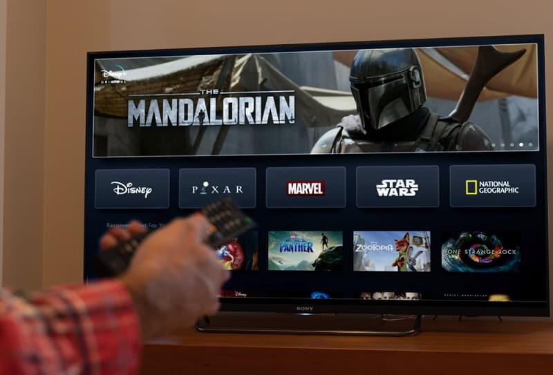 a hand holding TV remote to search for the Mandalorian on Disney+